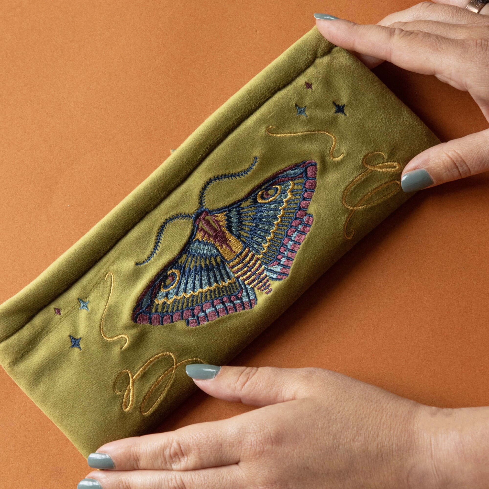 Green clutch with embroidered moth design held by hands on an orange background